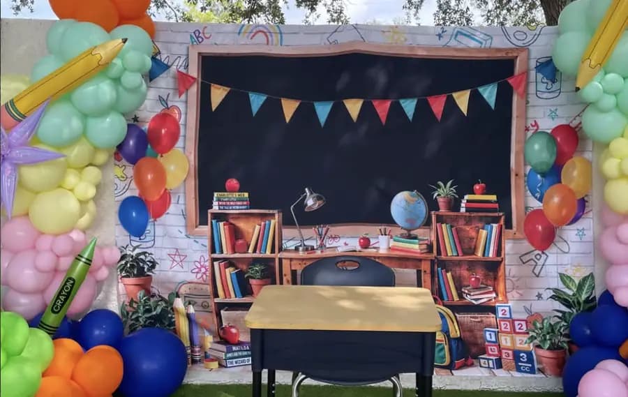 Un montaje de vuelta al cole con pizarra, pupitre y dos arcos de globos bajo un árbol.